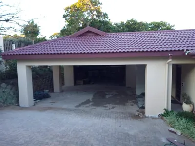 Open carport with maroon tiled roof, cream pillars, and paved concrete floor in landscaped KZN home.